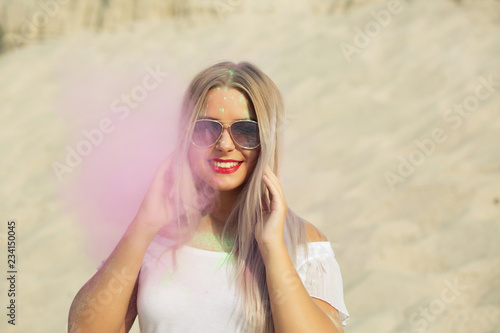 Emotional blonde girl wearing shirt with naked shoulders and glasses playing with pink dry paint Holi at the desert