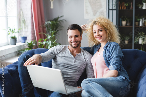 Couple searching locations a trip with laptop sitting on a couch in the living room at home.