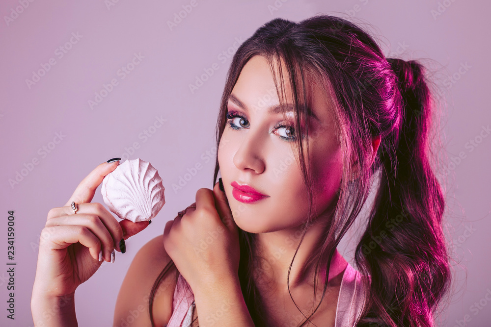 A glamorous woman on a pink-purple background holds a white marshmallow in her hand