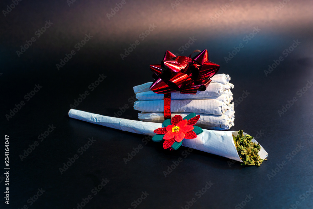 A large marijuana joint with a festive decoration on it in front of a bundle of joints tied in ribbon and a bow isolated.