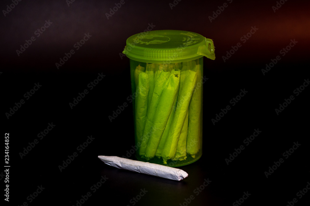 A bright transparent green RX pill bottle filled with marijuana joints with a single joint laying infront of it.