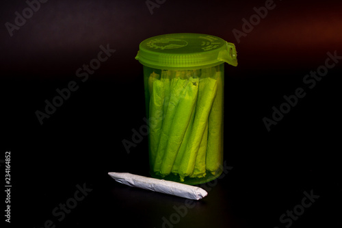 A bright transparent green RX pill bottle filled with marijuana joints with a single joint laying infront of it.