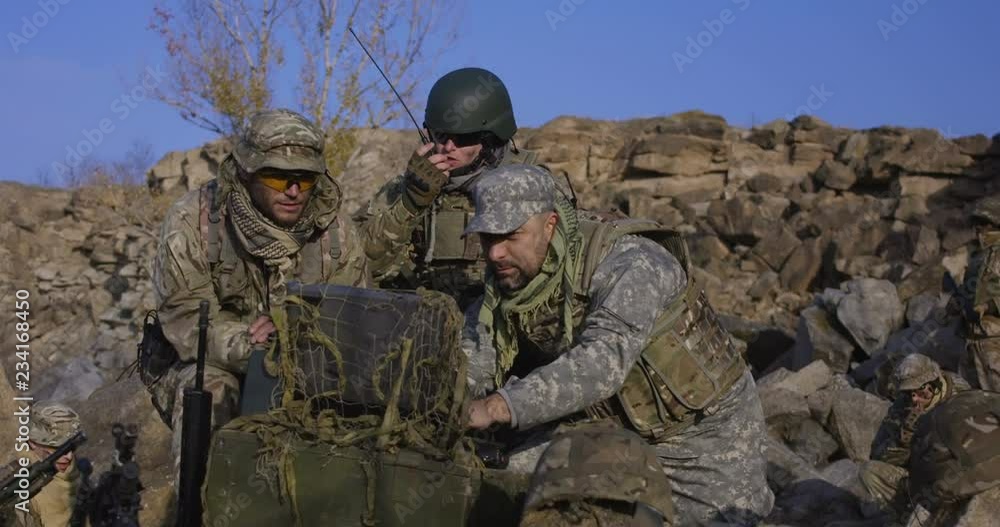 Medium shot of fully equipped and armed soldiers looking at a computer and talking on a radio in the field Medium shot of fully equipped and armed soldiers looking at a computer and talking on a radio in the field