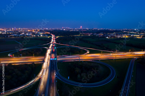Cloverleaf interchange seen from above. Aerial view of highway road junction in the night with light trails of evening traffic. Bird's eye view. City lights in the background.