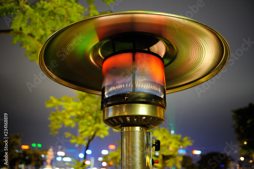Close up at radiant outdoor gas heater working at night in the street restaurant. Green-orange toned reflections. Tree and buildings in background.