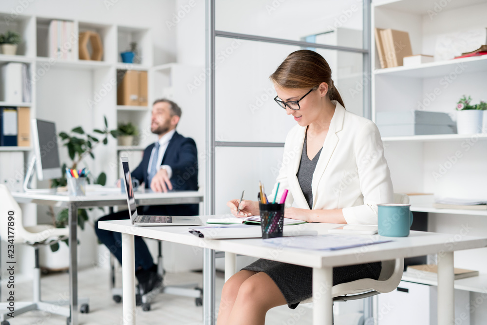 Content busy attractive lady in white jacket and eyeglasses sitting at table and making notes in diary while planning schedule in office Content busy attractive lady in white jacket and eyeglasses sitting at table and making notes in diary while planning schedule in office