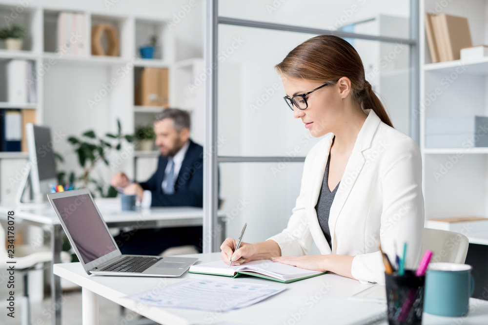 Serious busy young lady in glasses sitting at table with laptop and papers while making notes in diary and writing out ideas Serious busy young lady in glasses sitting at table with laptop and papers while making notes in diary and writing out ideas