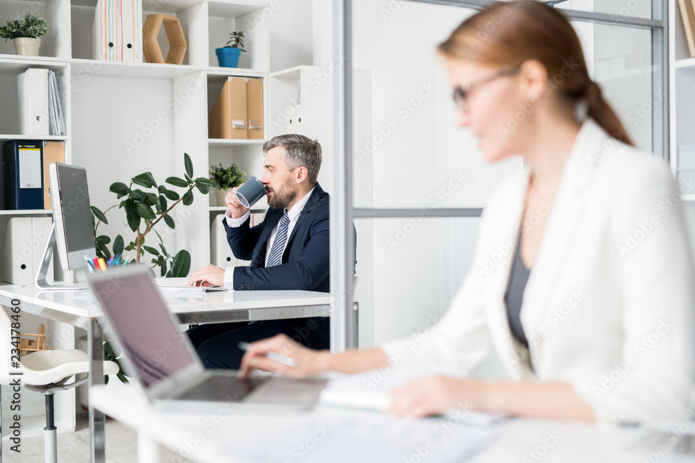 Serious pensive bearded businessman in formal jacket sitting at table and drinking coffee while using computer and working in office