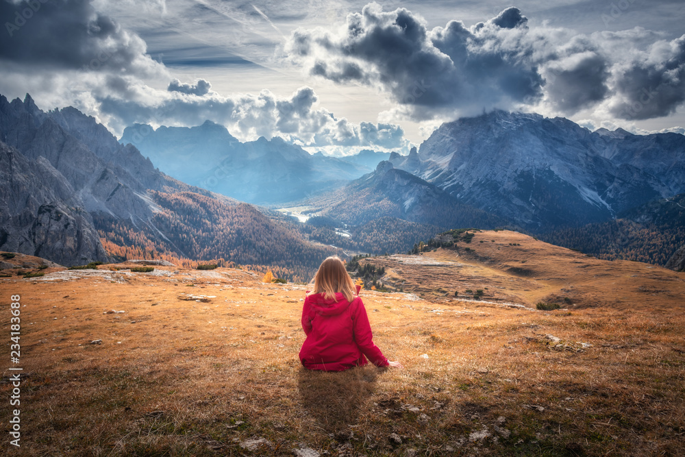 Fototapeta premium Young woman is sitting on the hill against the majestic mountains at sunset in autumn in Dolomites, Italy. Landscape with girl, cloudy sky, orange grass, high rocks with forest in fall. Italian alps