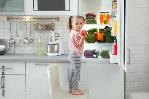 Cute girl taking apple out of refrigerator in kitchen