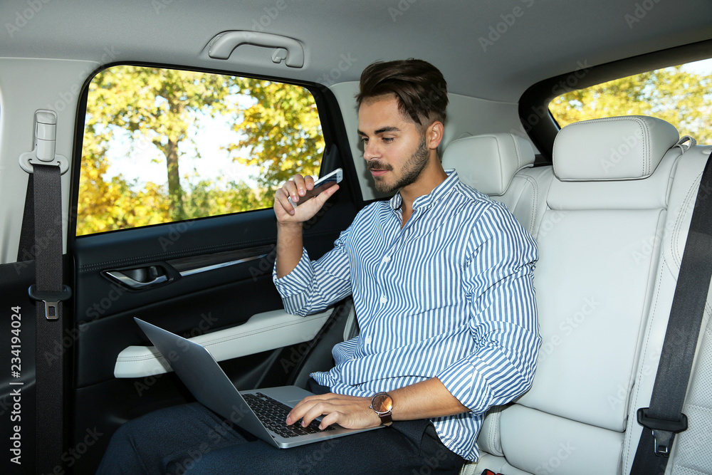 Young handsome man with phone using laptop in back seat of car