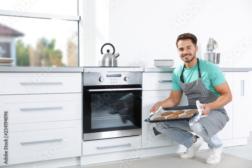 Young man holding baking sheet with cookies near oven in kitchen