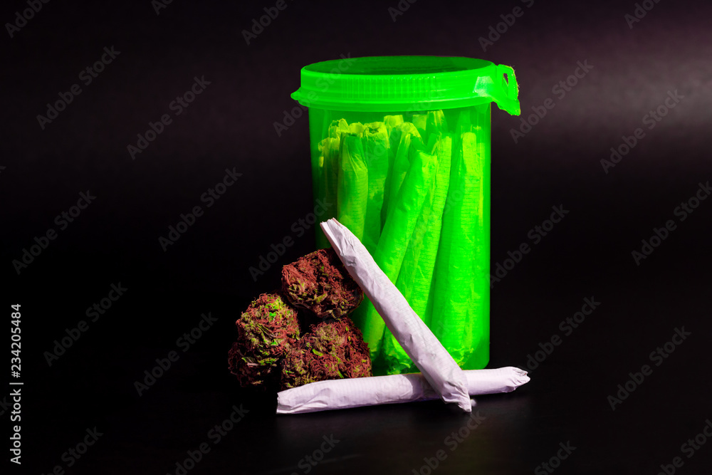 A bright transparent green RX pill bottle filled with marijuana joints next to a pile of buds with 2 joints.