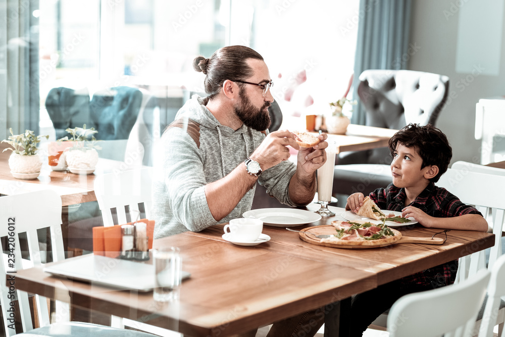 Lovely son. Beaming bearded man wearing glasses looking at his lovely son while eating pizza for lunch - obrazy, fototapety, plakaty 