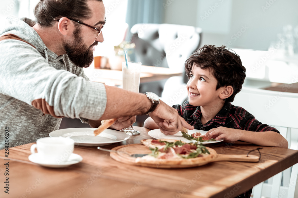 Tasting pizza. Bearded dark-haired father wearing glasses tasting cheesy pizza with his cute funny son Tasting pizza. Bearded dark-haired father wearing glasses tasting cheesy pizza with his cute funny son