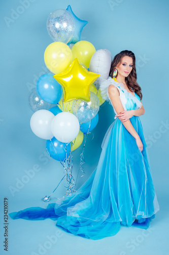 Happy girl in prom with helium air white, blue and yellow balloons. Portrait of a beautiful woman in a blue dress with a lot of colorful balloons studio blue background.concept freedom and celebration