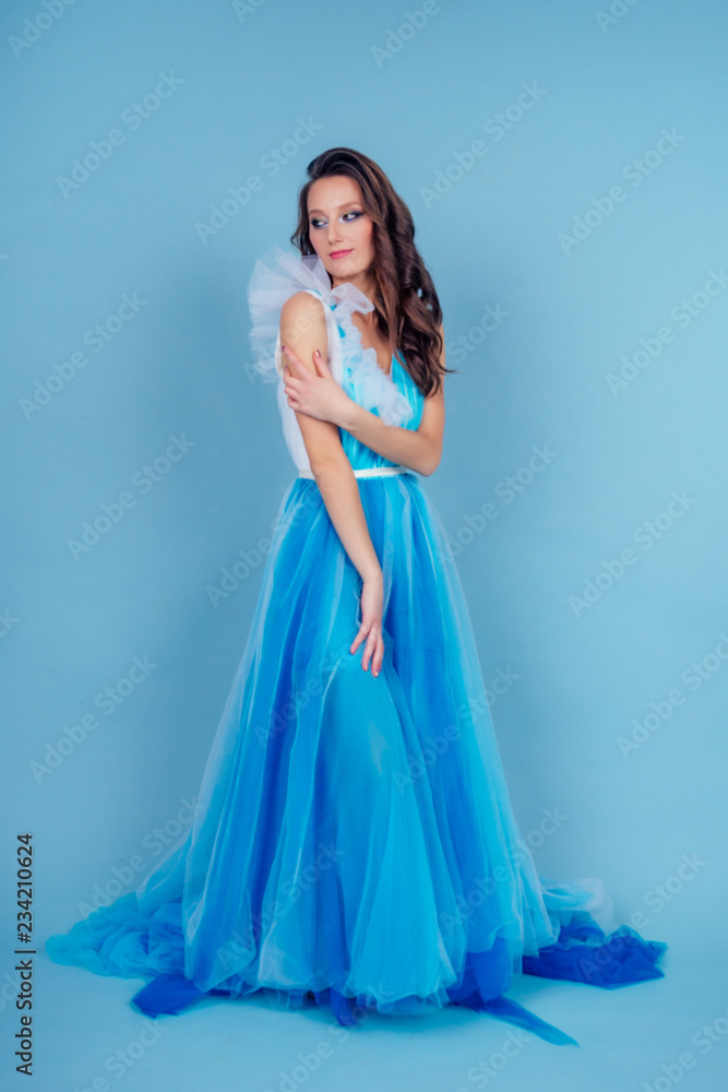 Young beauty woman in tulle fluttering blue dress.Beautiful girl with make-up , hairstyle and gorgeous look.female model prom seamstress and designer clothes tailor on a blue background in the studio Young beauty woman in tulle fluttering blue dress.Beautiful girl with make-up , hairstyle and gorgeous look.female model prom seamstress and designer clothes tailor on a blue background in the studio