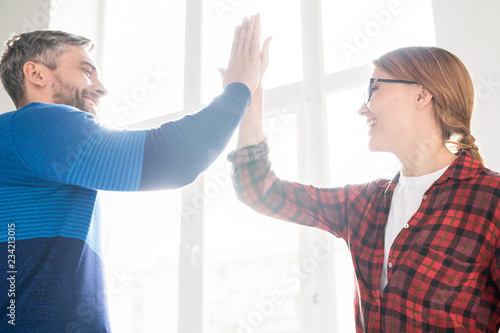 Cheerful successful young startuppers in casual clothing standing in modern office against window and giving high five in excitement photo