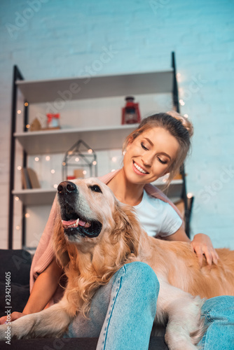beautiful cheerful young blonde woman sitting on couch and hugging golden retriever dog at christmas time