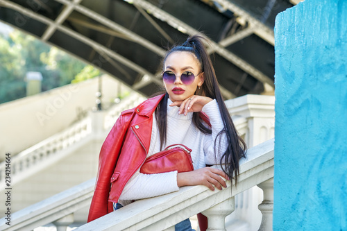 Portrait of a fashionable girl in a red leather jacket and sunglasses in the city