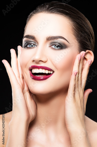 Young woman with expression emotion, bright red glossy lips make-up, smokey eye shadows, smile, clean skin, hands touhing face. Black background photo