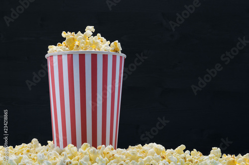A red-white paper cup filled with popcorn stands in grains of corn, on a dark background.