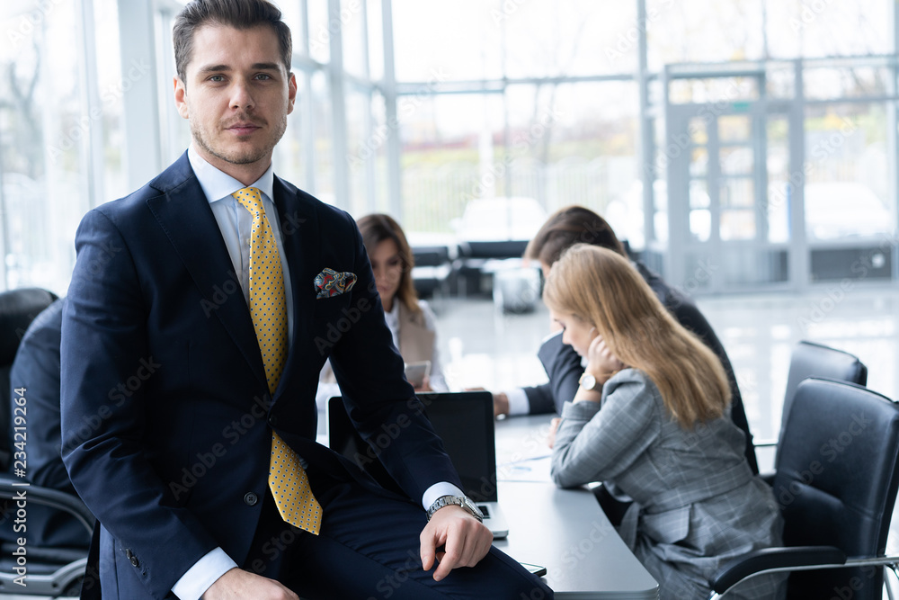 Young and confident businessman. Handsome young man in formalwear smiling at camera while his colleagues working on background. Young and confident businessman. Handsome young man in formalwear smiling at camera while his colleagues working on background.