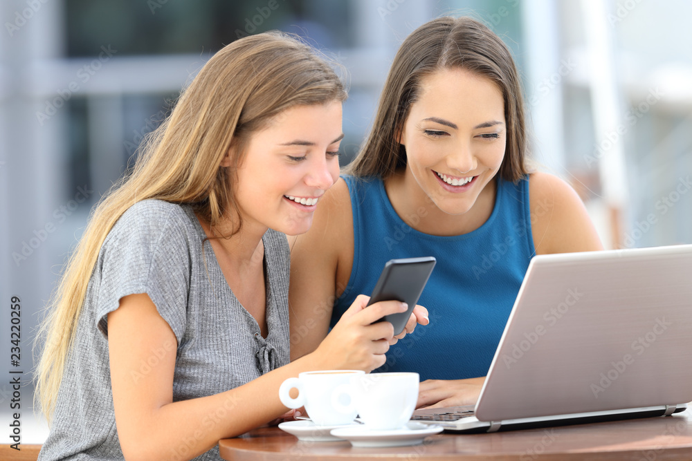 Two happy friends using laptop and phone in a bar Two happy friends using laptop and phone in a bar