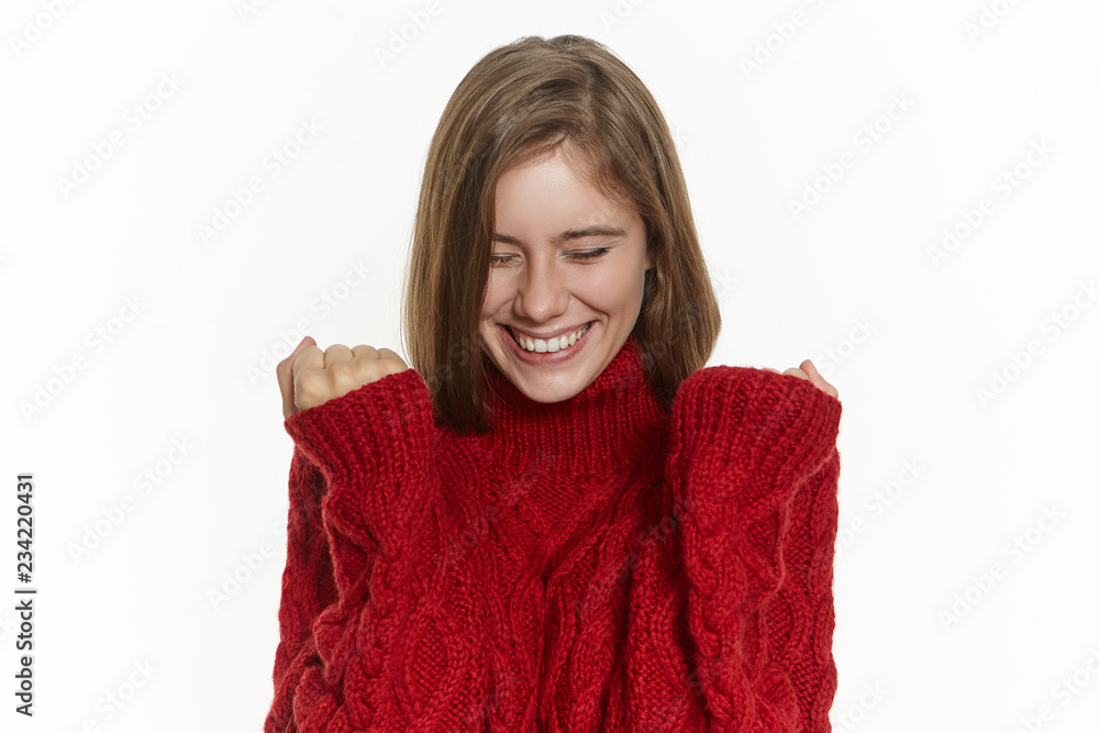 Success, joy and happiness concept. Happy excited young female in stylish warm sweater posing isolated keeping fists clenched and smiling broadly, rejoicing at good news, making her dream come true Success, joy and happiness concept. Happy excited young female in stylish warm sweater posing isolated keeping fists clenched and smiling broadly, rejoicing at good news, making her dream come true