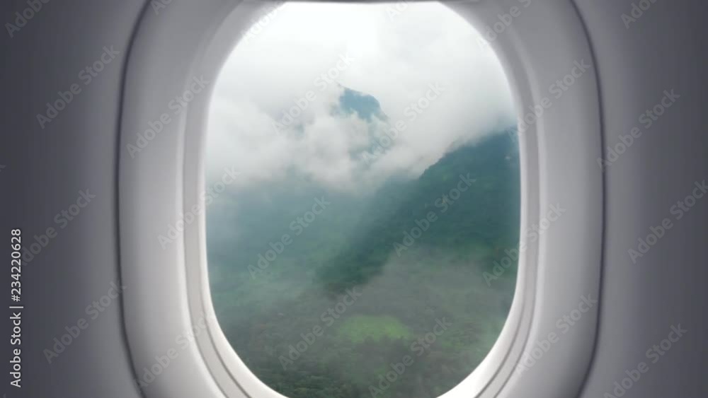 Zoom in view from the window at passenger seat in airplane flight above green mountain valley with clouds Zoom in view from the window at passenger seat in airplane flight above green mountain valley with clouds