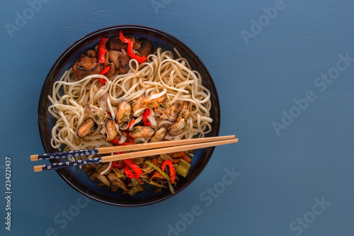 Noodles with seafood, salad, red pepper and fried mushrooms in a traditional porcelain plate on a blue table. Copy space. Top view