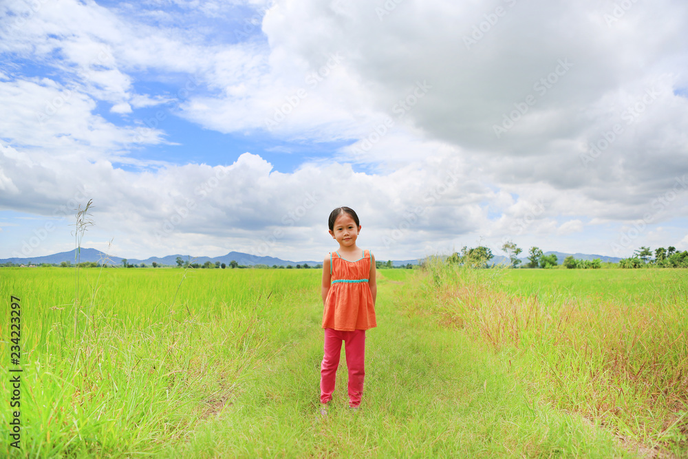 Smiling little Asian child girl standing in the young green paddy fields with mountain and cloud sky.