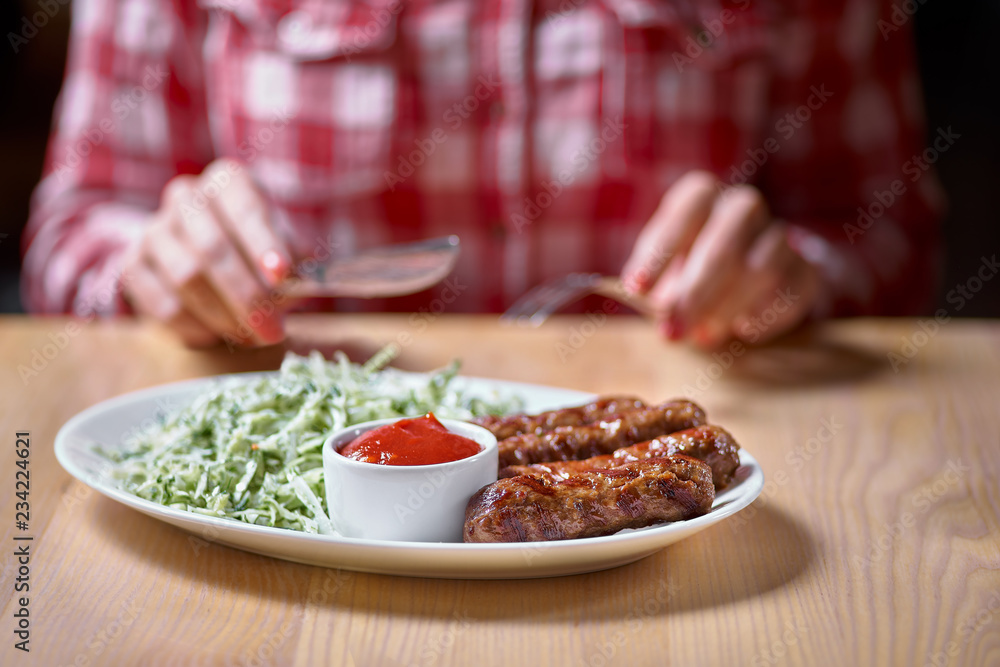 Woman cuts sausage grill with a fork and knife in a restaurant, in a red plaid shirt. selective focus.restaurant. copy space for design menu Woman cuts sausage grill with a fork and knife in a restaurant, in a red plaid shirt. selective focus.restaurant. copy space for design menu