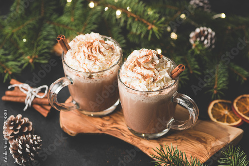 Two cups of hot chocolate with whipped cream and cinnamon on wooden serving board surrounded with fir tree, Christmas lights, cinnamon and pine cones. Cozy Christmas Drink