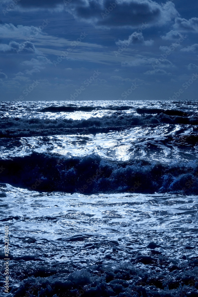 Fototapeta premium A blue sea glistening white with dark blue waves and a blue cloudy sky