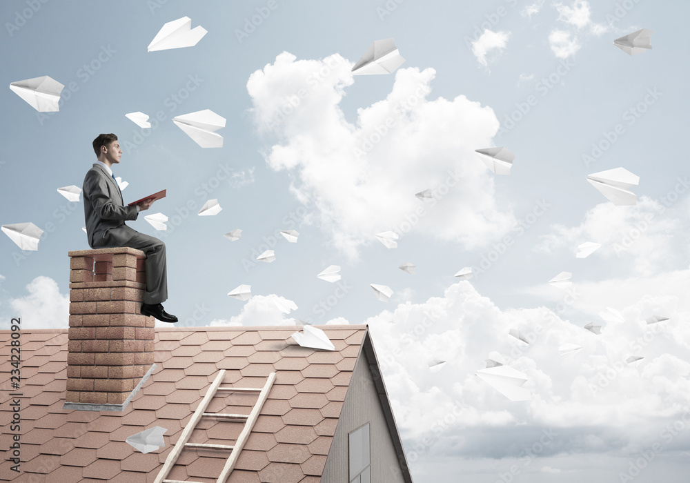 Man on brick roof reading book and paper planes flying in air Man on brick roof reading book and paper planes flying in air