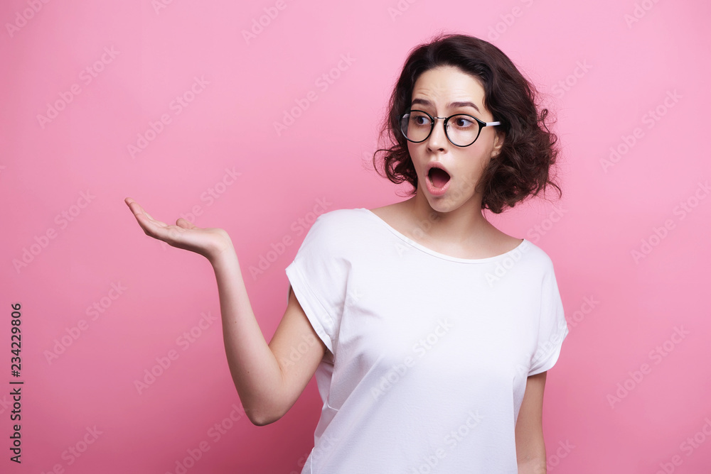 Good looking young Caucasian woman in round transparent eyewear, keeps hand raised, dressed in casual outfit, pretends holding something wonderful, isolated over pink background. Look there Good looking young Caucasian woman in round transparent eyewear, keeps hand raised, dressed in casual outfit, pretends holding something wonderful, isolated over pink background. Look there