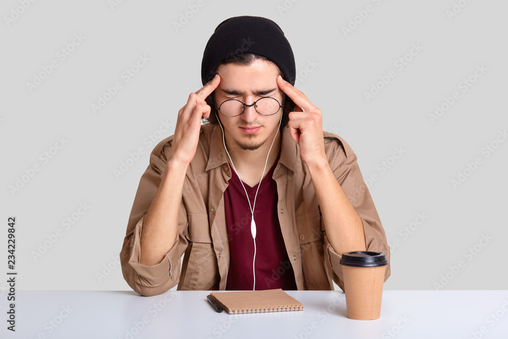 Horizontal shot of tired man keeps both index fingers on temples, tries to gather with thoughts, concentrate on something, closes eyes, dressed in hat, shirt, writes in notebook isolated on white wall Horizontal shot of tired man keeps both index fingers on temples, tries to gather with thoughts, concentrate on something, closes eyes, dressed in hat, shirt, writes in notebook isolated on white wall