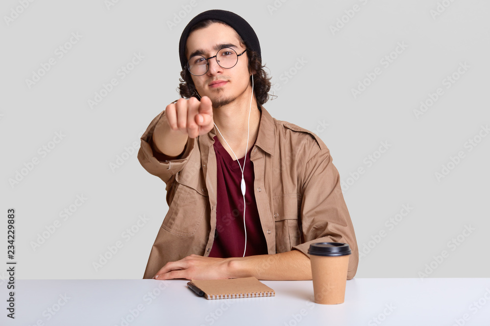 Image of attractive male wears round glasses, sits at desktop with coffee and organizer, points directly at camera, dressed in fashionable clothes, isolated over white background. You are chosen Image of attractive male wears round glasses, sits at desktop with coffee and organizer, points directly at camera, dressed in fashionable clothes, isolated over white background. You are chosen