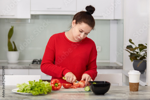 Horizontal shot of European woman waits for baby, cuts tomatoes, makes fresh salad, uses knife, poses against kitchen interior, had dark hair combed in knot, being vegetarian. Healthy eatting concept