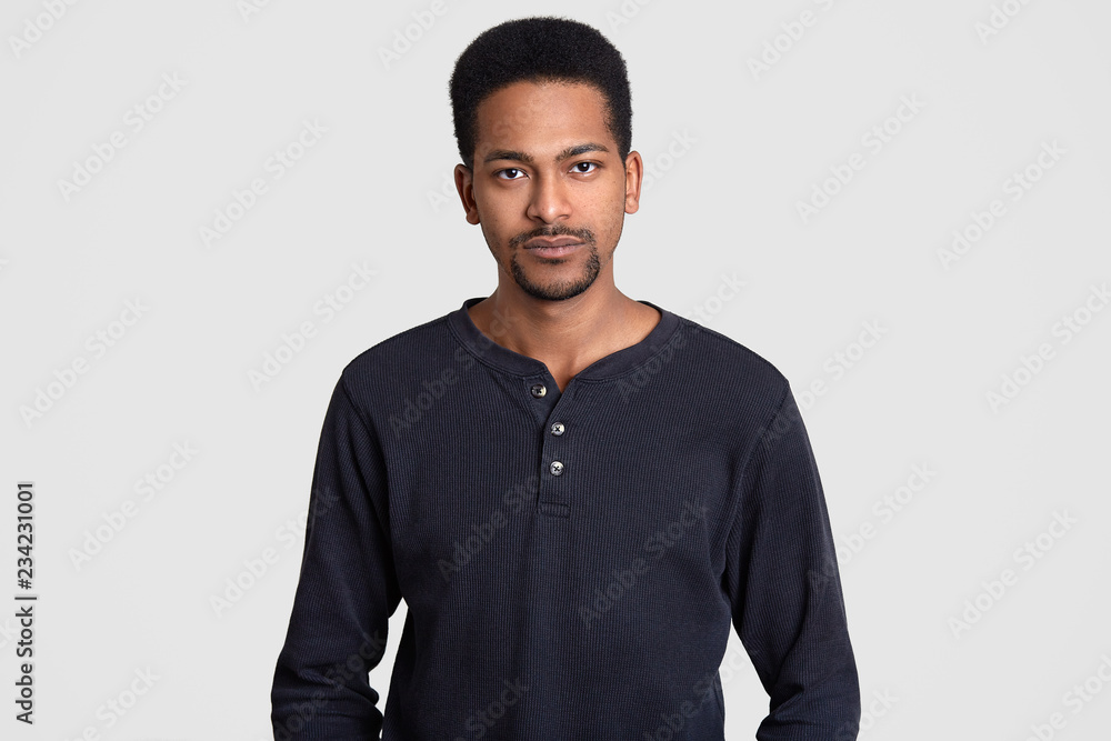 Serious Afro American man dressed in casual sweater, has self confident expression, looks directly at camera, isolated over white background. Ethnic male poses at studio. Facial expressions concept