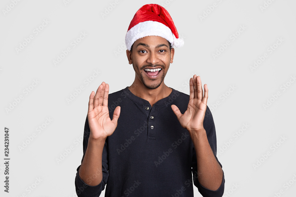 Glad black man raises hands and shows palms with happiness, wears Santa Claus hat, casual jumper, smiles broadly, shows perfect white teeth, models in studio. New Year and ethnicity concept. Glad black man raises hands and shows palms with happiness, wears Santa Claus hat, casual jumper, smiles broadly, shows perfect white teeth, models in studio. New Year and ethnicity concept.