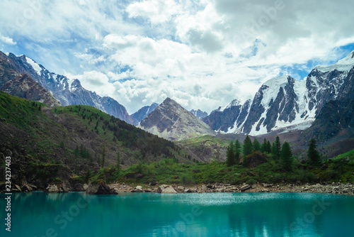 Mountain lake on front of giant beautiful glacier. Amazing mountain in shape of pyramid. Snowy ridge under cloudy sky. Wonderful atmospheric landscape of majestic nature. Coniferous trees in highlands