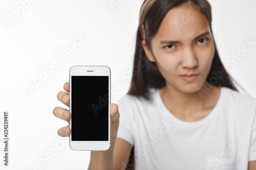 Modern technology and communication concept. Serious confident young Korean woman holding generic smart phone with black blank copyspace screen for your advertisement. Selective focus on gadget