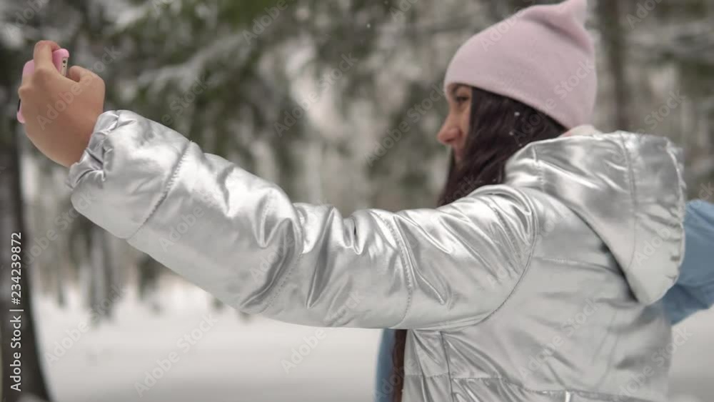 Two young girlfriends in winter clothes taking selfie against the background of the winter forest using a smartphone and smiling. Two young girlfriends in winter clothes taking selfie against the background of the winter forest using a smartphone and smiling.