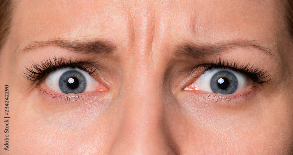 Naklejka premium Close-up przestraszona twarz pięknej młodej kobiety o pięknych niebieskich oczach i dużych ładnych rzęsach i brwiach. Makro oczu ludzkich - zdziwienie lub szok, patrząc w kamerę.