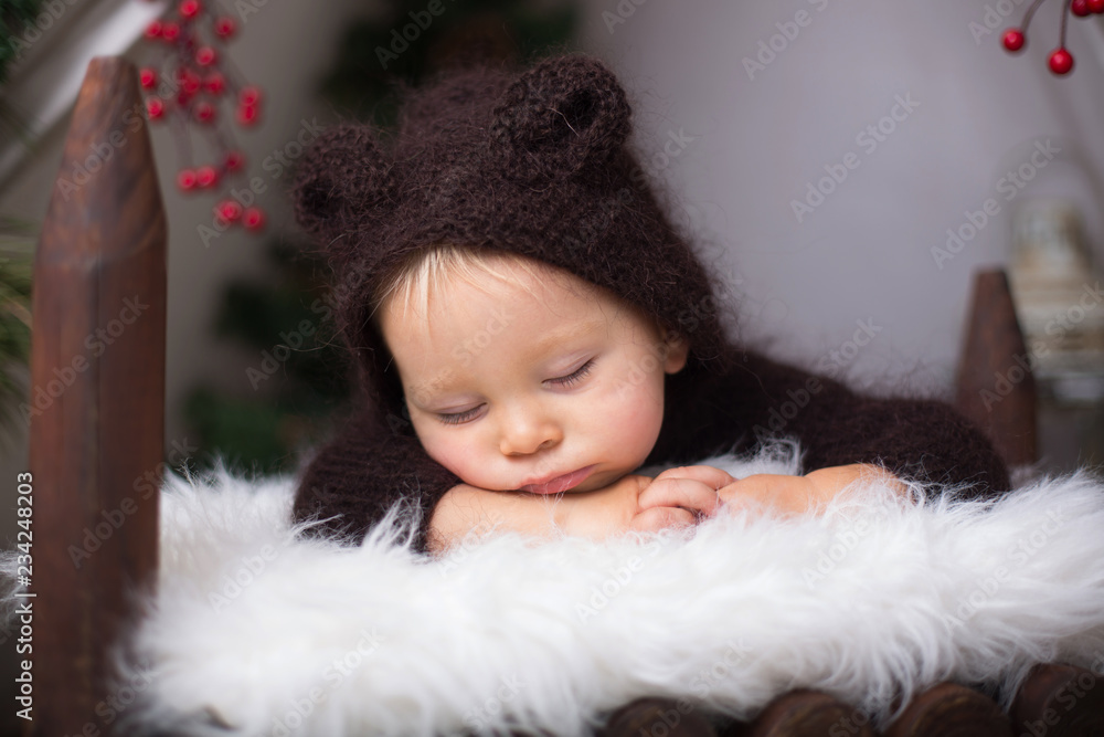 Cute toddler baby boy in handmade knitted bear costume, sleeping in wooden bed in teepee,  Christmas setup