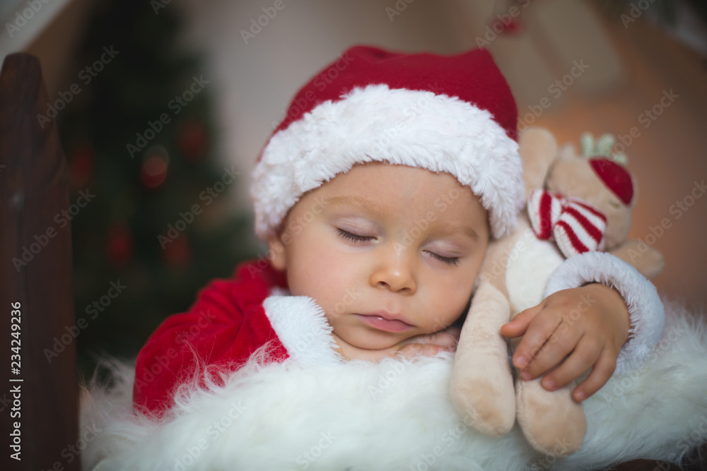 Adorable little toddler baby boy dressed in canta claus costume, sleeping in baby bed in front of crhistmas teepee Adorable little toddler baby boy dressed in canta claus costume, sleeping in baby bed in front of crhistmas teepee