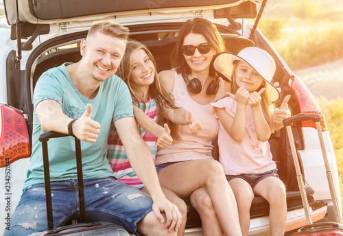 Happy pleasured family sitting on a trunk of a car with suitcases on a travel vacation