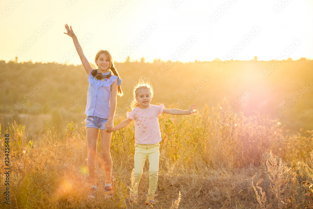 Little smiling girls stand on sunshine evening field with joyfully raised hands - obrazy, fototapety, plakaty 